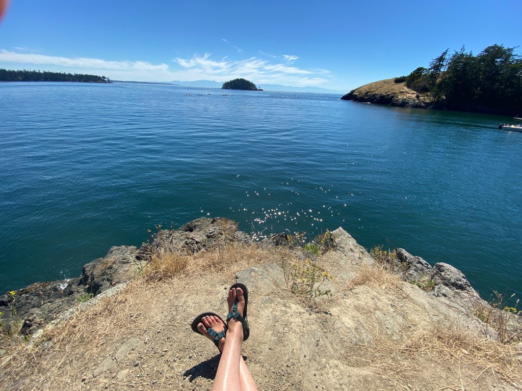 justine fanarof's legs with bedrock thong sandals sitting on an outcrop overlooking the water of deception pass state park in washington state usa islands in the distance heartspace